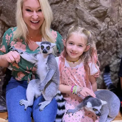 A smiling mother and daughter with ring-tailed lemurs climbing on them.